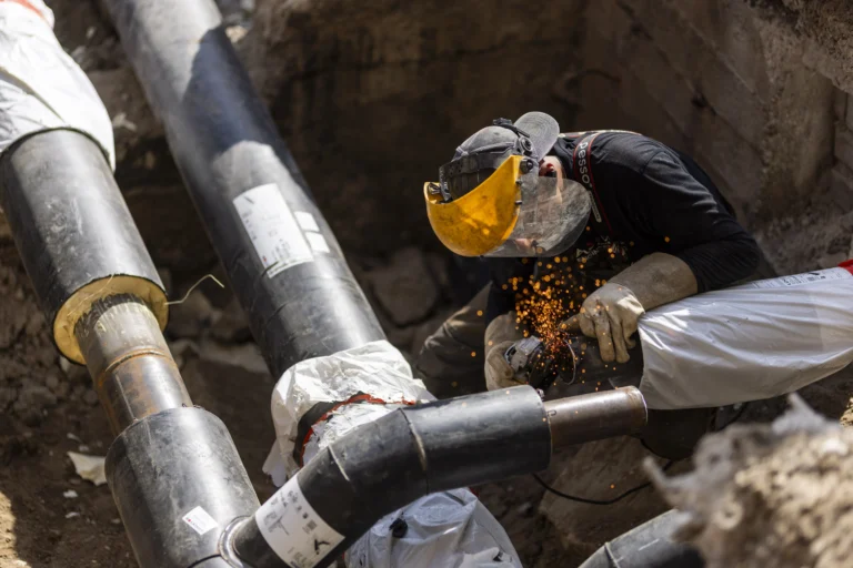 Construction worker wearing a face shield grinds metal pipes, producing bright sparks in an excavation trench.