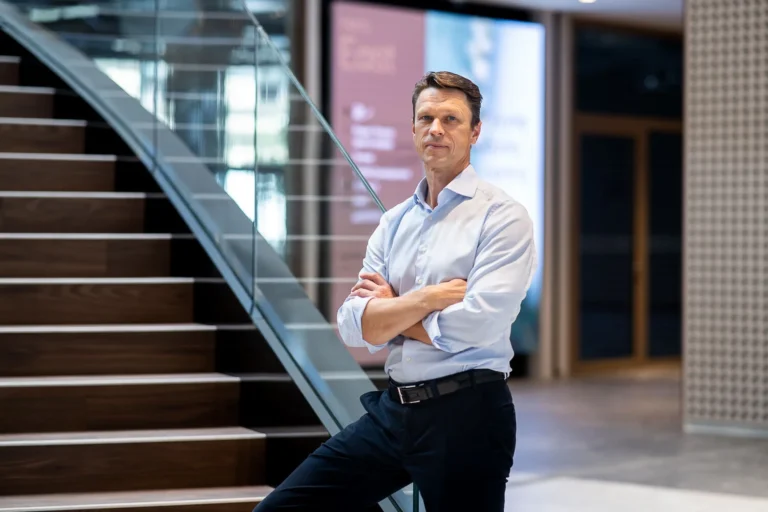 Man with crossed arms in a light blue shirt stands by a modern office staircase.