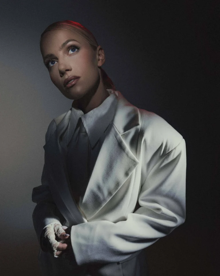 Blonde woman wearing an oversized white blazer and lace gloves looking up in dramatic lighting.