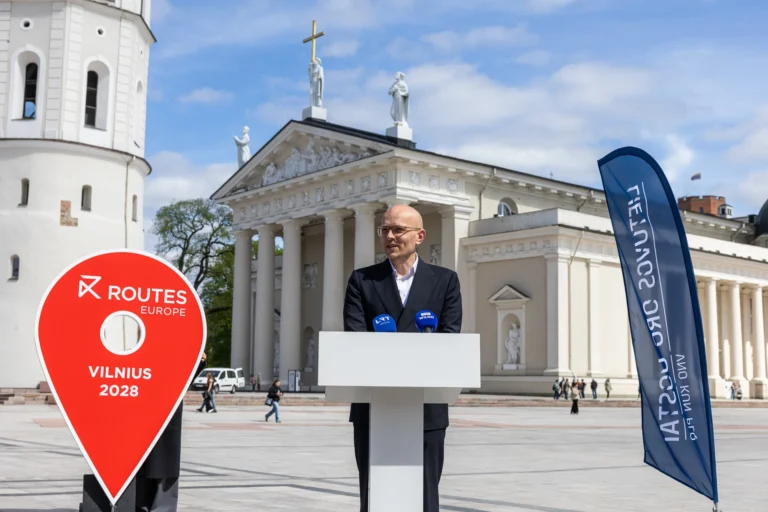 Man speaking at a podium before Vilnius Cathedral with a large red location pin sign.
