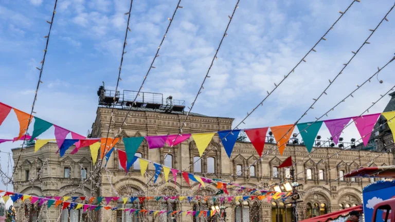 Colorful festive bunting flags strung across a historic city square with an ornate stone building in the background under a blue sky.