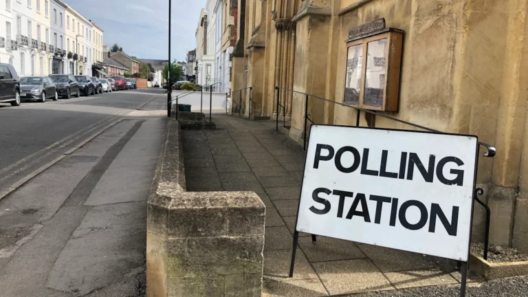 A white 'Polling Station' sign stands on a pavement outside a traditional building on a residential street.