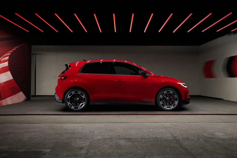 Side profile of a red electric hatchback parked in a studio with red neon ceiling lights.