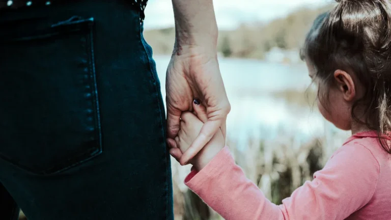 A close-up of a foster parent holding a young child's hand while looking out over a body of water.