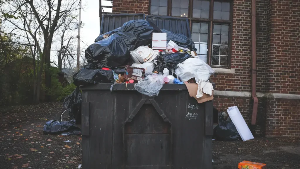 An overflowing industrial waste bin outside a brick building in a urban setting.