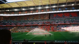 Football supporters watching from the stands of a large stadium as sprinklers water the pitch.