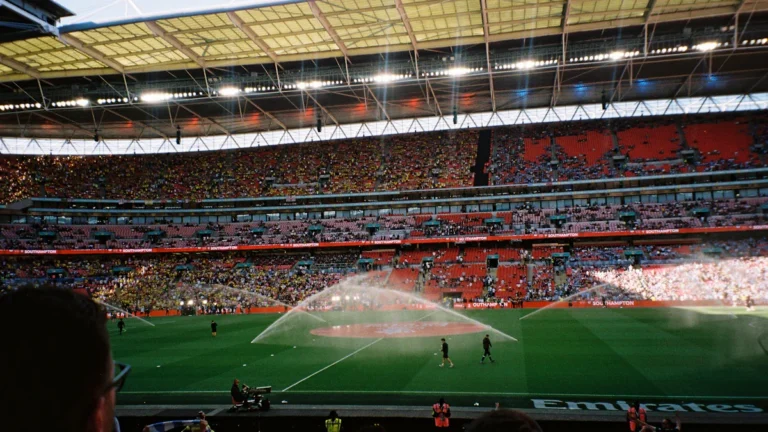 Football supporters watching from the stands of a large stadium as sprinklers water the pitch.