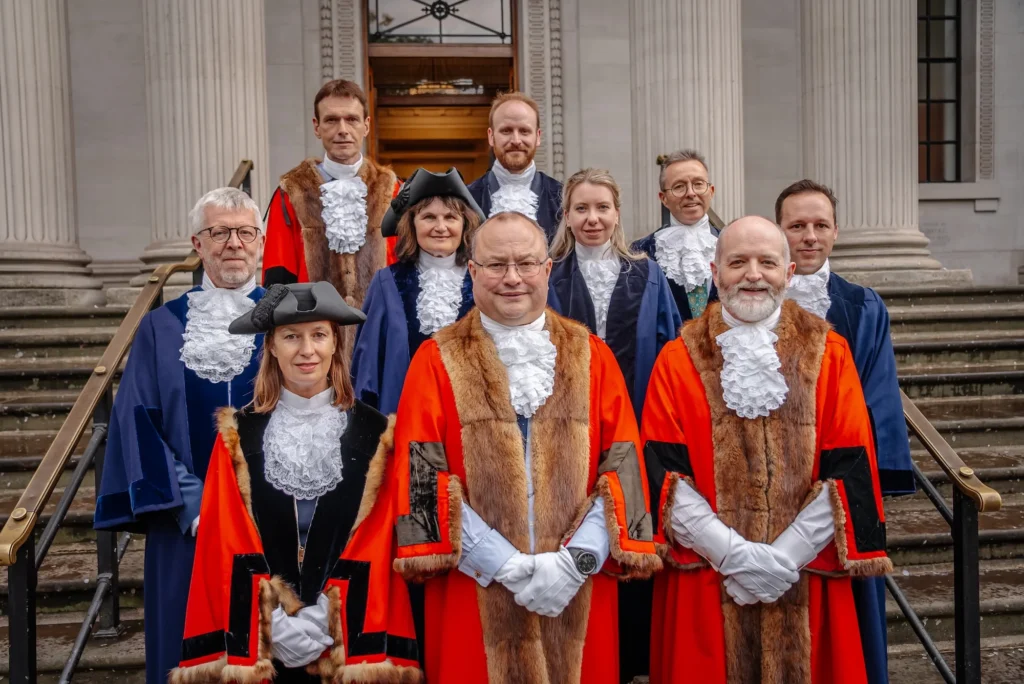 Officials wearing traditional red and blue ceremonial robes with white lace jabots stand on building steps.