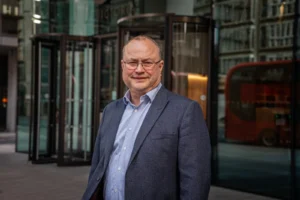 A man with glasses wearing a blue blazer and light blue shirt outside a glass building.