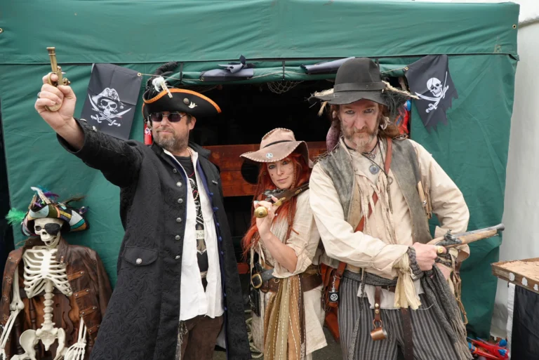 Three people in pirate costumes pose with replica pistols in front of a green tent.