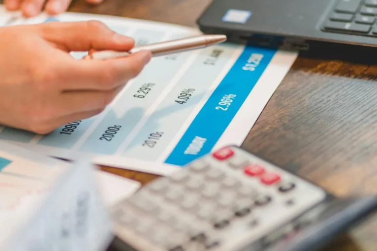 Hand pointing a pen at financial documents showing percentage data next to a calculator and laptop.