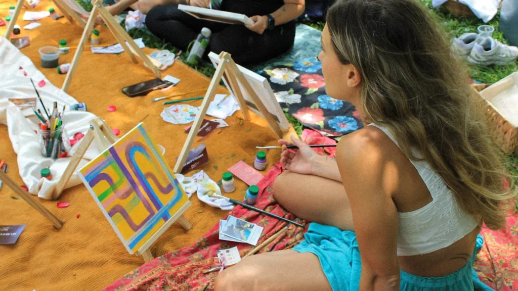 A group of people sitting on blankets outdoors, painting on small canvases with easels as part of a community art project.