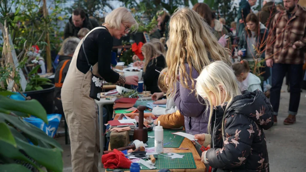 A diverse group of Wigan residents, including children and adults, participating in a community arts and crafts workshop in a bright, plant-filled space.