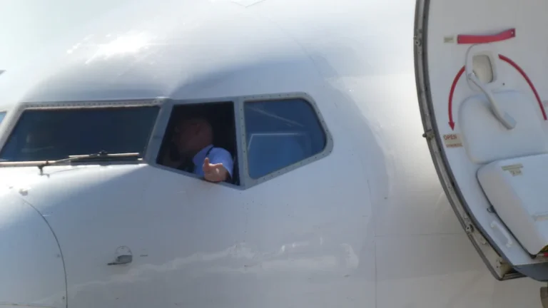 A pilot in a white uniform shirt looks out from the cockpit window of a commercial airplane.