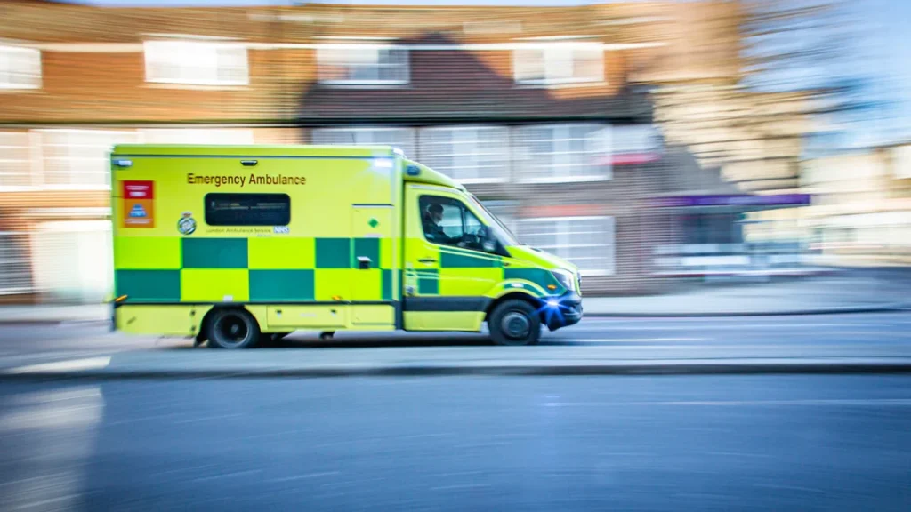 An emergency ambulance van with high-visibility markings traveling along a UK road.