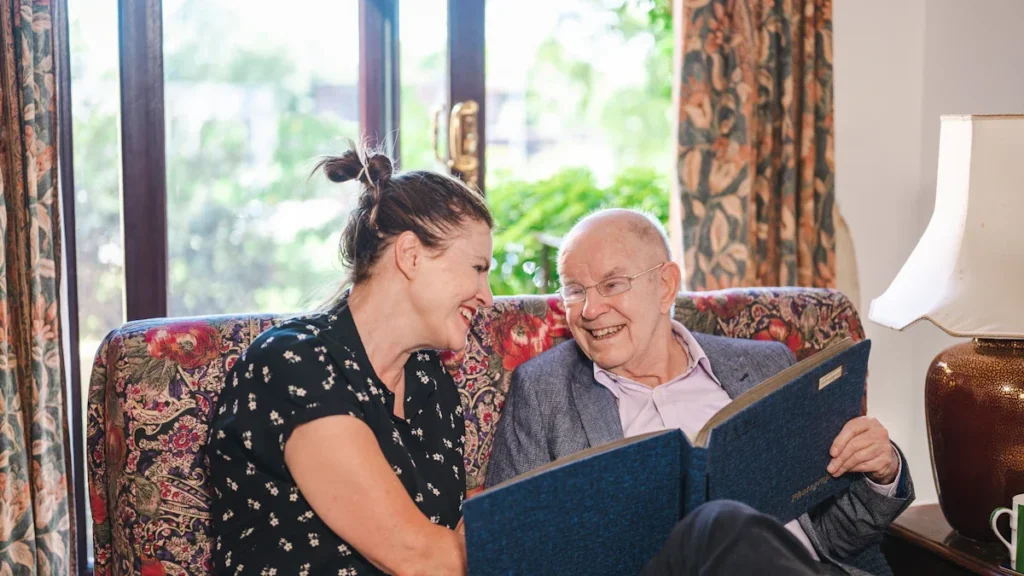 A friendly caregiver reading a book with an older man on a sofa.