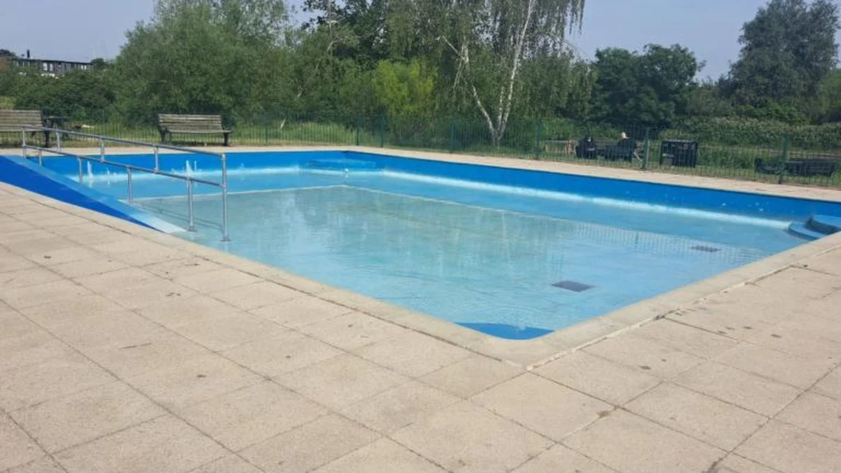 An empty outdoor swimming pool with blue tiled edges and a metal accessibility ramp.