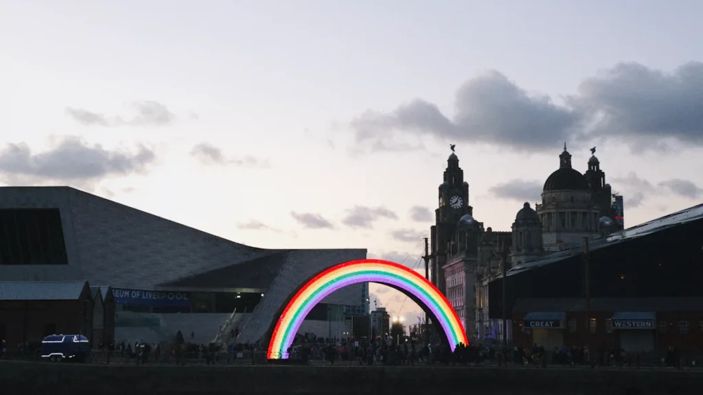 A glowing rainbow arch installation located in front of Liverpool's historic waterfront buildings.