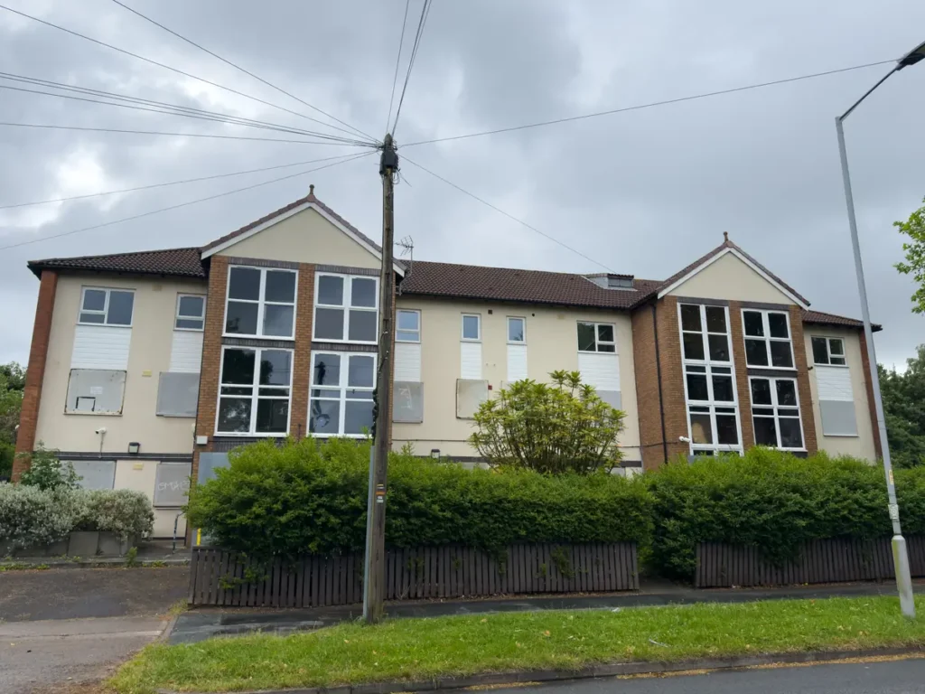 A two-story residential building with boarded-up windows behind a low wooden fence.