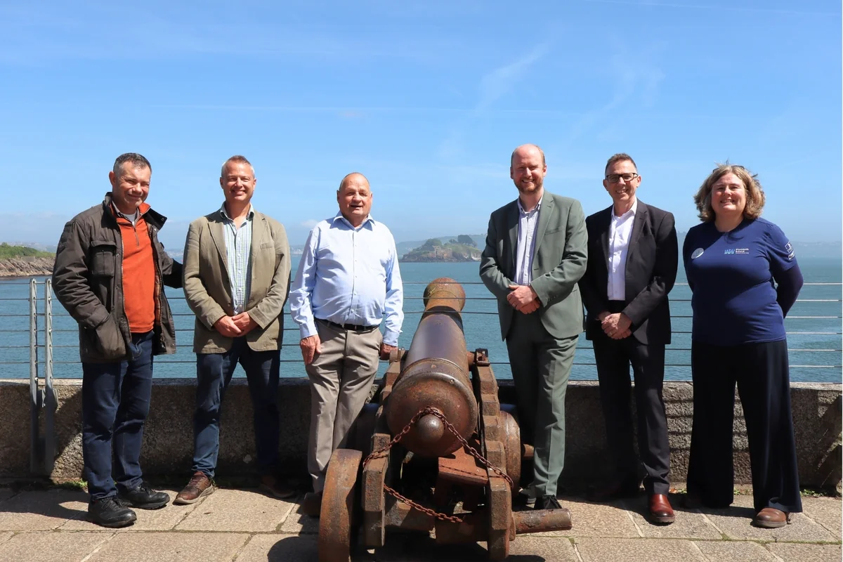 Group of six adults standing together outdoors behind a historic iron cannon by the sea.