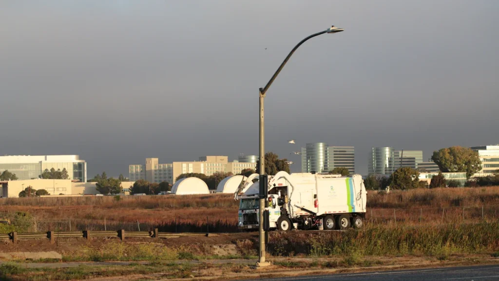 A white waste collection vehicle driving along a road near office buildings.