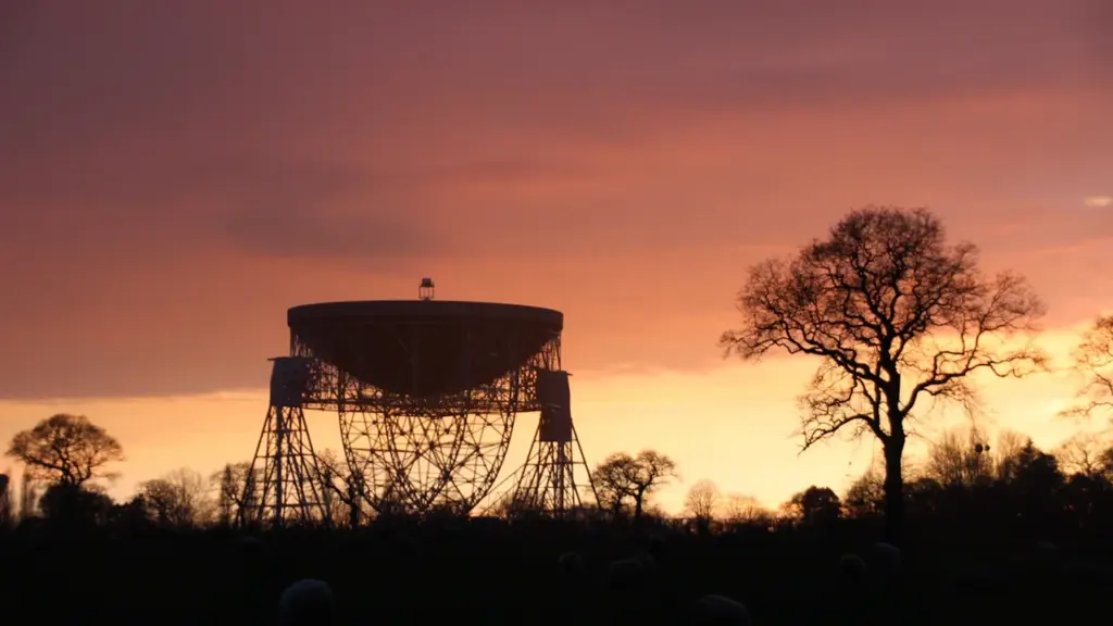 The iconic Lovell Telescope at Jodrell Bank silhouetted against a vibrant orange sunset.
