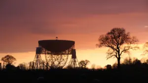 The iconic Lovell Telescope at Jodrell Bank silhouetted against a vibrant orange sunset.