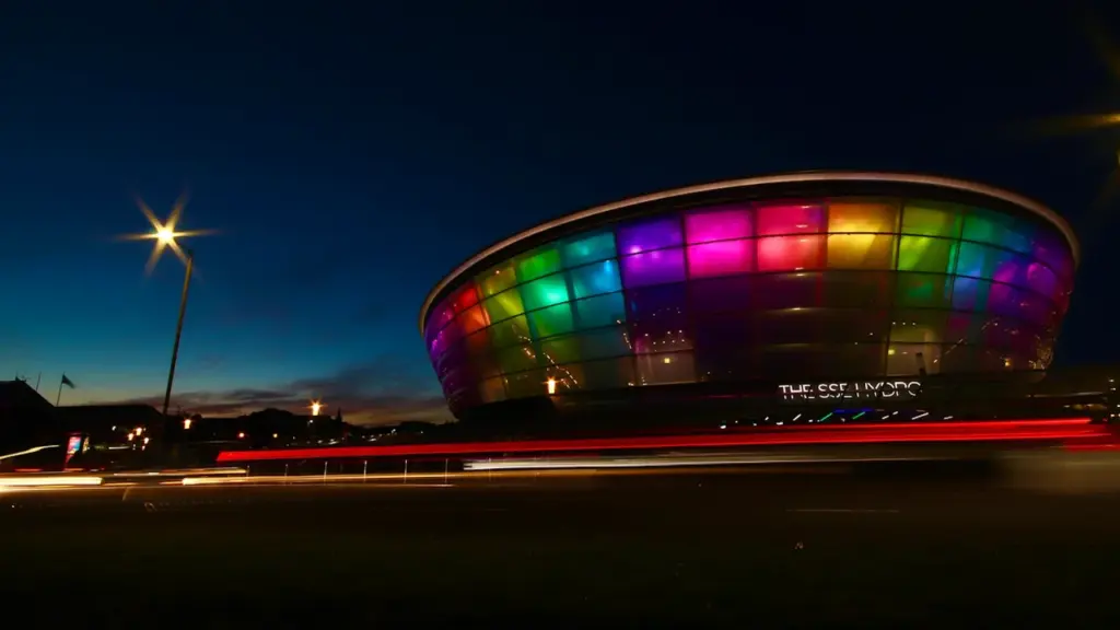 The OVO Hydro arena in Glasgow illuminated at night with vibrant multi-colored lights.
