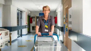 A volunteer pushing a trolley through a clean, modern UK hospital corridor.