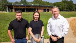 Three people stand outdoors in a sunny park before a modern brick building.