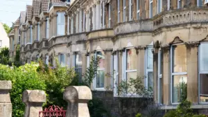 A row of historic Victorian terraced stone houses with bay windows and lush front gardens.