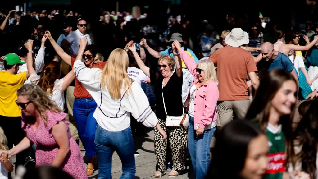 A diverse crowd of people dancing and holding hands in a bright outdoor square.