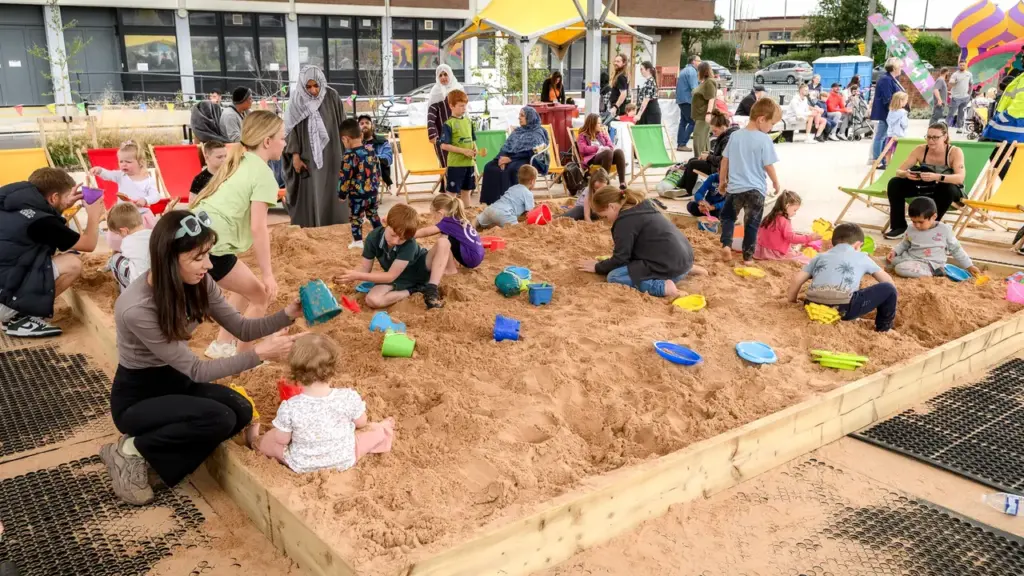 Many children and parents play together in a large outdoor wooden sandbox with toys.