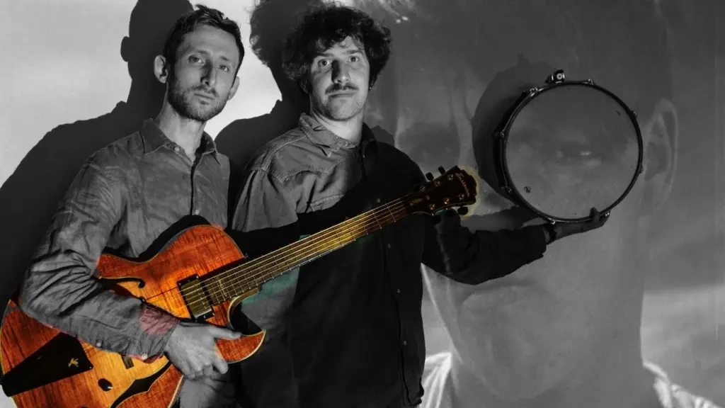 Two men pose, one holding an electric guitar and the other holding a snare drum.