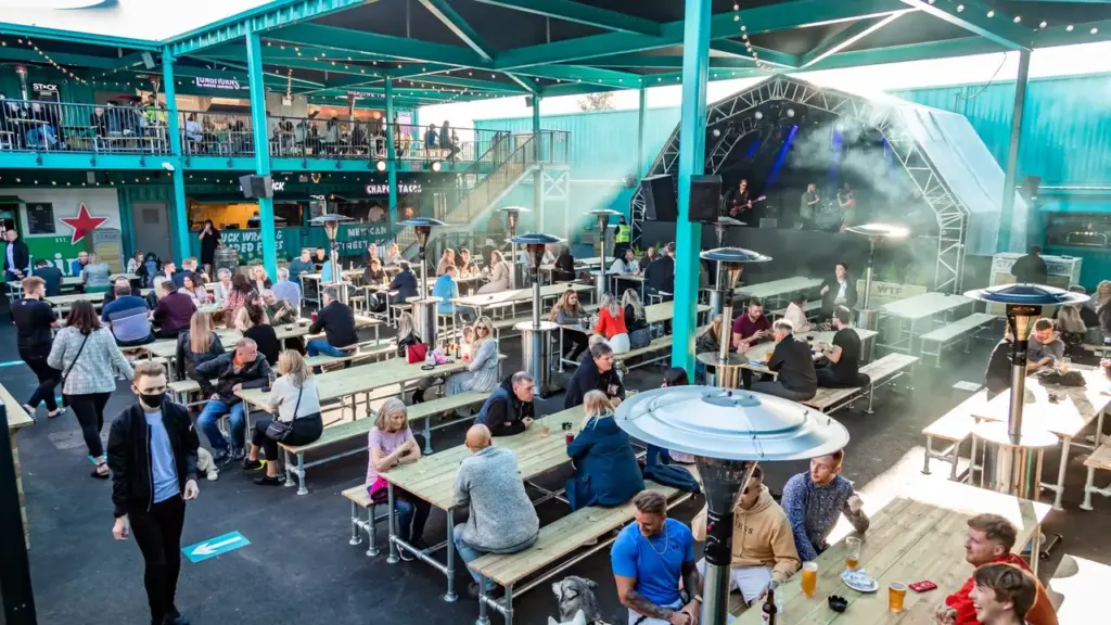 People sit at outdoor picnic tables in a vibrant container-themed leisure venue with live music.