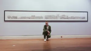 A man in a green suit crouches before a long, detailed architectural sketch of buildings.