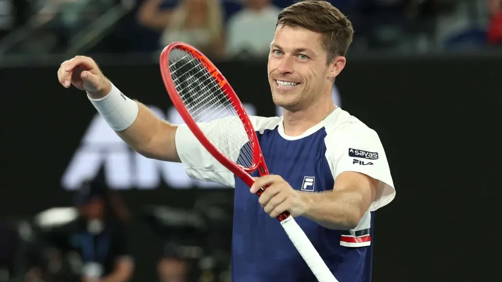 A smiling male tennis player holds a red tennis racket on a professional court.