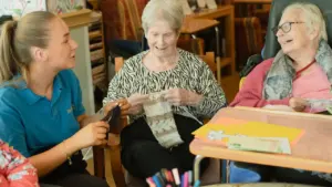Caregiver assisting two elderly women with knitting in a Welsh care facility.