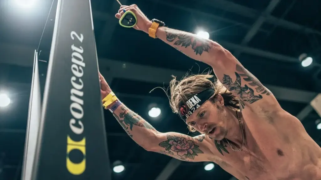 A tattooed man with a headband uses a Concept2 SkiErg exercise machine intensely.