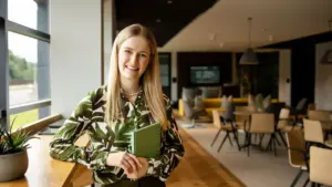 A smiling blonde woman wearing a green leaf-patterned blouse holds a notebook in an office.