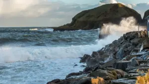 Strong waves crash against a rocky coastline under a cloudy, late afternoon sky.
